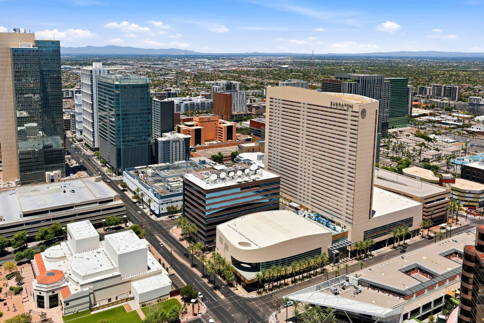 An aerial of the Phoenix Convention Center’s curved roof and the surrounding downtown street grid, with mountains in the distance